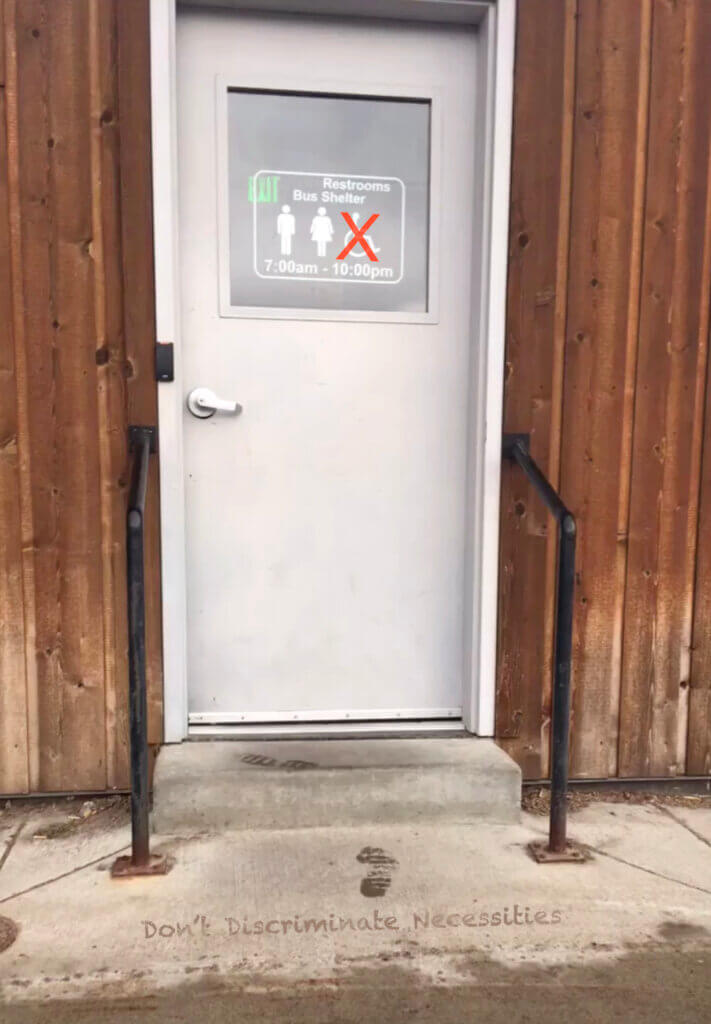 A white door with black handle bars on the sides surrounded by a wooden exterior. The door has a window with printed restroom icons of a male, a female, and the handicap symbol crossed out. In front of the door is a cement step with the phrase “Don’t Discriminate Necessities” written in a font that looks like water in front of the step.