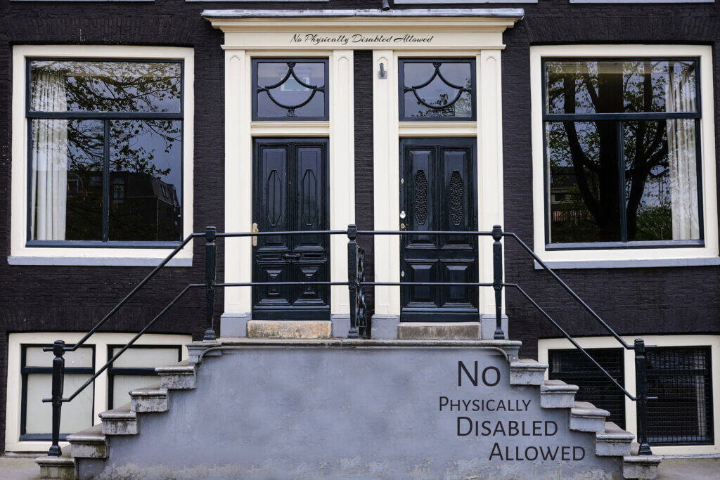 A dark navy apartment duplex double doorway with a staircase on the right leading up to it. The phrase “No Physically Disabled Allowed” is written under each step to the left side. On the center white trim above the doorways is the same phrase written also.